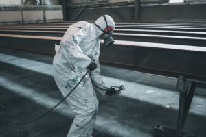 A worker in protective gear using a sandblasting hose to clean a large steel surface outdoors.