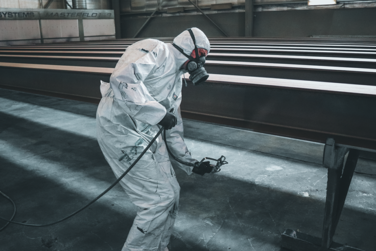 A worker in protective gear using a sandblasting hose to clean a large steel surface outdoors.